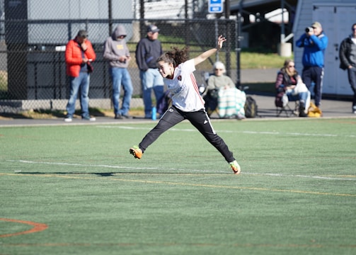 A person is mid-air on a sports field, appearing to be involved in an athletic activity. Several spectators are on the sidelines, some seated and others standing, with one individual capturing the event with a camera. The background includes a fence and various casual observers, some of whom are wearing winter clothing.
