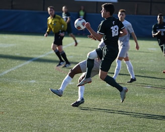 A soccer match taking place on a grassy field. Players are in action, with one player in black jersey number 18 controlling the ball mid-air while being challenged by an opposing player in a white jersey. A referee in a yellow uniform observes the game closely in the background, along with other players.