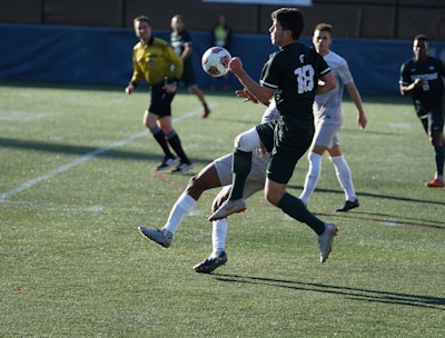 A soccer match taking place on a grassy field. Players are in action, with one player in black jersey number 18 controlling the ball mid-air while being challenged by an opposing player in a white jersey. A referee in a yellow uniform observes the game closely in the background, along with other players.