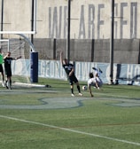 A soccer match on a field where players are in action near a goalpost. Several players in black uniforms are positioned in and around the goal area, while one player in white is executing an acrobatic move mid-air with a soccer ball nearby. The backdrop includes a tall wall with faded writing on it, and the field features green artificial turf with blue markings.