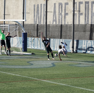 A soccer match on a field where players are in action near a goalpost. Several players in black uniforms are positioned in and around the goal area, while one player in white is executing an acrobatic move mid-air with a soccer ball nearby. The backdrop includes a tall wall with faded writing on it, and the field features green artificial turf with blue markings.