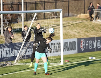 A soccer match scene with two players in focus near the goal. One player, wearing black, is in mid-air reaching to stop or catch the ball, while the other player, wearing teal shorts and black top, is positioned under the ball, looking up at it. The goal net and a fence are visible in the background, with a few spectators standing near the sidelines.