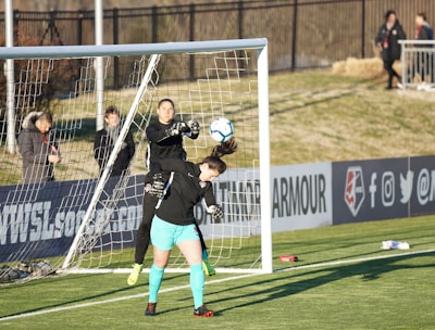 A soccer match scene with two players in focus near the goal. One player, wearing black, is in mid-air reaching to stop or catch the ball, while the other player, wearing teal shorts and black top, is positioned under the ball, looking up at it. The goal net and a fence are visible in the background, with a few spectators standing near the sidelines.