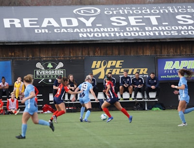 Players engage in a competitive soccer game on a field while others are seated on the bench watching attentively. The action is taking place in front of a wooden structure displaying multiple advertisements, including orthodontics, lacrosse, and field hockey.