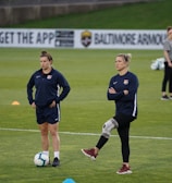 Two individuals are standing on a grassy sports field, each wearing dark athletic clothing with a logo. One person is balancing on one leg with a brace on their knee, while the other stands with hands on hips. Soccer balls and markers are visible in the background, along with a person collecting equipment.