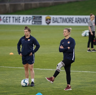 Two individuals are standing on a grassy sports field, each wearing dark athletic clothing with a logo. One person is balancing on one leg with a brace on their knee, while the other stands with hands on hips. Soccer balls and markers are visible in the background, along with a person collecting equipment.