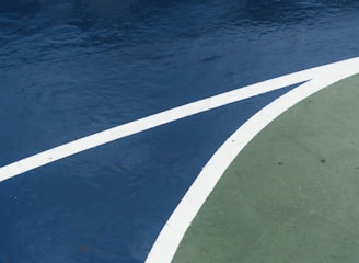 A close-up view of a sports court featuring distinctly marked lines, with a dark blue surface contrasted by a curve of green. The white lines delineate the boundary between the two areas, creating an abstract visual effect.