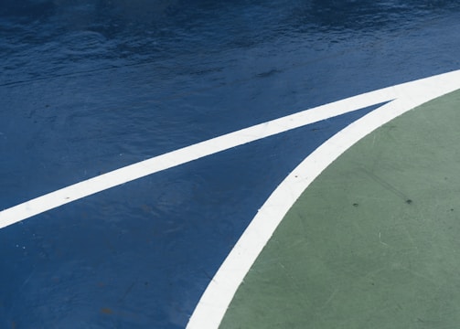 A close-up view of a sports court featuring distinctly marked lines, with a dark blue surface contrasted by a curve of green. The white lines delineate the boundary between the two areas, creating an abstract visual effect.