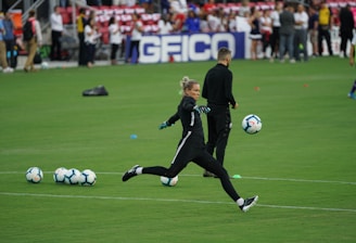 A soccer player in a black sports outfit is mid-action on a green field, focusing intently on kicking a soccer ball. Several soccer balls are scattered on the grass nearby, and a coach or teammate stands in the background watching. Spectators can be seen in the distant stands.