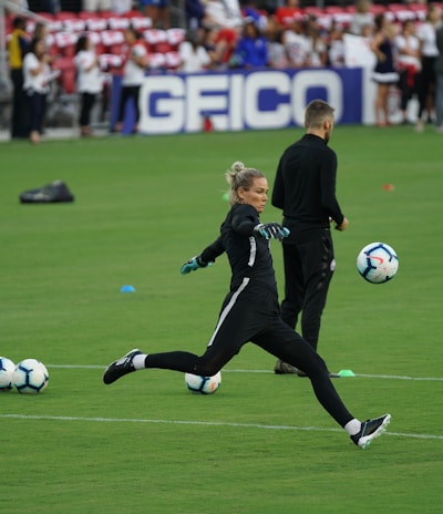 A soccer player in a black sports outfit is mid-action on a green field, focusing intently on kicking a soccer ball. Several soccer balls are scattered on the grass nearby, and a coach or teammate stands in the background watching. Spectators can be seen in the distant stands.