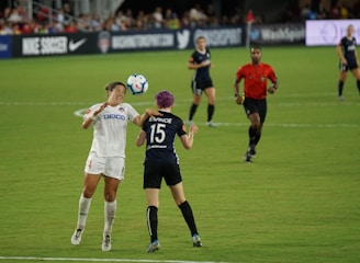 Two soccer players are engaged in a match on a grassy field, with one player attempting to head the ball while an opponent positions herself nearby. A referee in a red shirt observes the game along with other players in the background. Advertising banners and a crowd of spectators are visible along the sidelines.