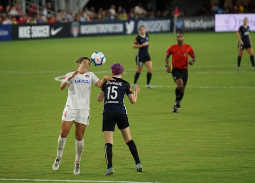 Two soccer players are engaged in a match on a grassy field, with one player attempting to head the ball while an opponent positions herself nearby. A referee in a red shirt observes the game along with other players in the background. Advertising banners and a crowd of spectators are visible along the sidelines.