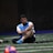 A man sits on a sports field, wearing a blue sports vest over a white shirt and black shorts. His legs are crossed, and he appears to be resting or taking a break. The background is dimly lit, with a blurred out soccer ball in the foreground and netting visible behind him.