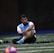 A man sits on a sports field, wearing a blue sports vest over a white shirt and black shorts. His legs are crossed, and he appears to be resting or taking a break. The background is dimly lit, with a blurred out soccer ball in the foreground and netting visible behind him.