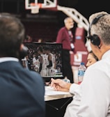 Two men wearing headsets focus on a monitor displaying a basketball team in discussion. Others can be seen around them, including a woman with a bottle of water, and an ongoing basketball game in the background.