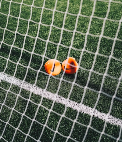 A close-up view of a football pitch, with a focus on two bright orange markers near the goal net. The green artificial turf is marked with a white line, and the net adds a texture to the scene.