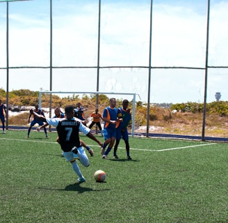 A soccer match is taking place on a field with players actively engaged in the game. One player wearing a black and white jersey with the number 7 is about to kick the ball. Opponents in blue and orange bibs are positioned near the goal, and a referee dressed in red observes the action from the side.
