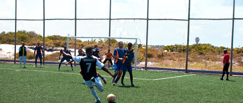 A soccer match is taking place on a field with players actively engaged in the game. One player wearing a black and white jersey with the number 7 is about to kick the ball. Opponents in blue and orange bibs are positioned near the goal, and a referee dressed in red observes the action from the side.