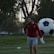 A man in sports attire stands on a soccer field, near a goalpost. He is wearing a red long-sleeve shirt and gray shorts, with a focused or pensive expression. Behind him, a large soccer ball is leaning against the net of the goal.