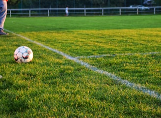 A soccer ball rests on a well-manicured grass field, bordered by fresh white lines marking the corner area. In the background, there are blurred figures and fencing, suggesting a sporting environment. One person's legs in jeans and sneakers are visible near the ball.