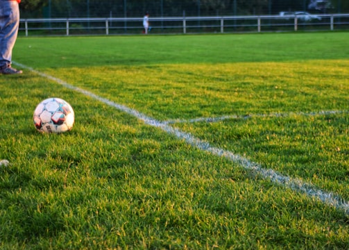 A soccer ball rests on a well-manicured grass field, bordered by fresh white lines marking the corner area. In the background, there are blurred figures and fencing, suggesting a sporting environment. One person's legs in jeans and sneakers are visible near the ball.