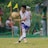 A soccer player in a white and blue uniform is seen in action on the field, focusing intently on controlling the ball with precision. Behind him, spectators watch from the sidelines, seated or standing near a net barrier. The setting indicates an outdoor event on a grassy pitch with vibrant green surroundings.