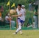 A soccer player in a white and blue uniform is seen in action on the field, focusing intently on controlling the ball with precision. Behind him, spectators watch from the sidelines, seated or standing near a net barrier. The setting indicates an outdoor event on a grassy pitch with vibrant green surroundings.